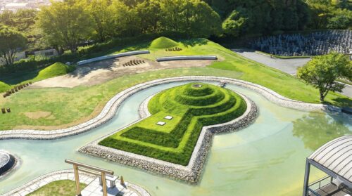 前方後円墳の墓所をリアルなイラスト調で描いた画像。堀の水、芝生の丘、鳥居、背景の木々や墓地が詳細に描かれている。