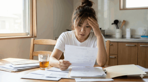 キッチンテーブルで書類の山を見つめ、困惑して額に手を当てている40代後半の女性